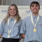 Christiansen and Often Take Gold - Carroll News | Carroll Publications | Carroll County, MD Two smiling SkillsUSA competitors wearing blue shirts with gold medals on stage beside a first-place sign.