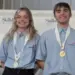Two smiling SkillsUSA competitors wearing blue shirts with gold medals on stage beside a first-place sign.
