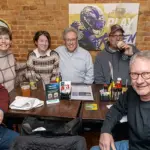 Trivia Team Serras - Carroll News | Carroll Publications | Carroll County, MD Group of seven adults smiling and seated around a wooden table in a brick-walled sports bar, Ravens logos on the wall, drinks and condiments on the table.