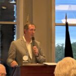 1778975966526-image - Carroll News | Carroll Publications | Carroll County, MD Man in a gray blazer speaks into a handheld microphone at a wooden podium during a meeting, with audience heads visible in the foreground and large windows showing a blue sky outside.