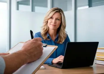 Businesswoman sits at a desk with a laptop, listening as a colleague takes notes on a clipboard in a modern office.