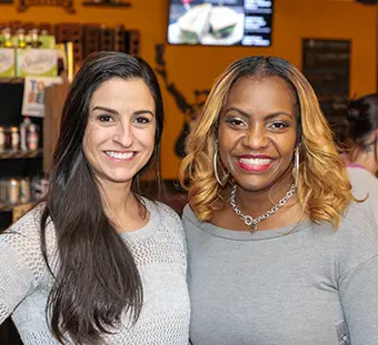 Two women posing for a photo in a casual indoor setting, smiling at the camera; one has long dark hair and a light gray knit sweater, the other has blonde hair and a gray top with a silver necklace.