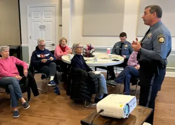 Senior adults seated at round tables in a community room, listening to a uniformed police officer giving a talk.