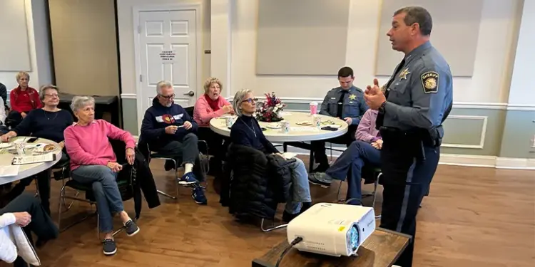 Financial Literacy Seminar_Featured Image - Carroll News | Carroll Publications | Carroll County, MD Senior adults seated at round tables in a community room, listening to a uniformed police officer giving a talk.
