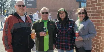 Four adults standing together outdoors at a community event, each holding a drink and smiling.
