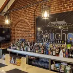 Interior_Bar - Carroll News | Carroll Publications | Carroll County, MD Brick-walled bar with wooden beams, a long white counter filled with assorted liquor bottles and a large Venue Bouchon sign on the brick wall behind.