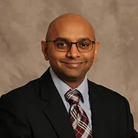 Professional headshot of a bald man in a dark suit, white shirt, and striped tie, wearing glasses and a subtle smile against a neutral backdrop.