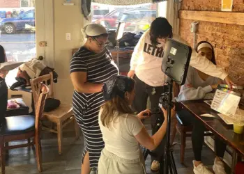 Group of people filming a video in a cafe, with a camera on a tripod and a monitor setup in the center of the scene.