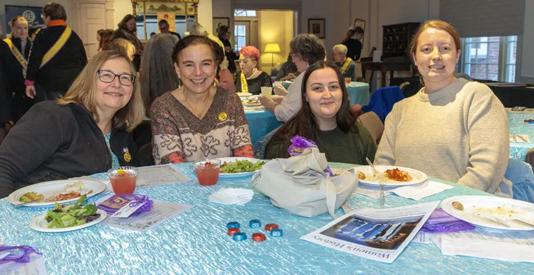 Four women sit at a table with a blue tablecloth, smiling at the camera during a community meal event at a busy gathering.