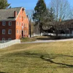 Union Mills Panorama - Carroll News | Carroll Publications | Carroll County, MD Brick building with a gabled roof and red doors along a street, white picket fences, and bare trees on a sunny day in a small town.