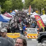 flower-jazz-fest-2026 - Carroll News | Carroll Publications | Carroll County, MD Crowded street festival with numerous vendor tents, American flags, and a road-closed barrier stretching along the avenue.