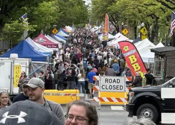 Crowded street festival with numerous vendor tents, American flags, and a road-closed barrier stretching along the avenue.