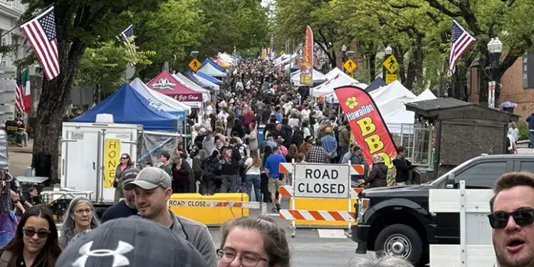 flower-jazz-fest-2026 - Carroll News | Carroll Publications | Carroll County, MD Crowded street festival with numerous vendor tents, American flags, and a road-closed barrier stretching along the avenue.
