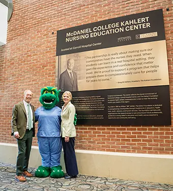 Two adults in business attire stand with a green dragon mascot beside a large informational plaque mounted on a brick wall that reads McDaniel College Kahler Nursing Education Center and related text.