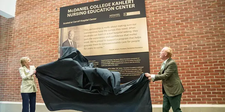 Two people unveil a plaque for the McDaniel College Kahler Nursing Education Center against a brick wall outside a hospital setting.
