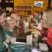mommy-date-image - Carroll News | Carroll Publications | Carroll County, MD Two women sit across from each other at a long communal table in a lively restaurant, toasting with drinks and smiling.