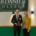 run-moore-mcdaniel - Carroll News | Carroll Publications | Carroll County, MD Two adults stand on a stage under a McDaniel College banner; man in a suit holds an award plaque, woman in a yellow blazer smiles beside him.