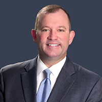 Smiling man in a dark suit and light blue tie against a dark blue background.