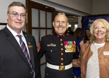 Three smiling adults at a formal event: a Marine in dress uniform with medals flanked by a man in a suit and a woman in a tan jacket nearby.