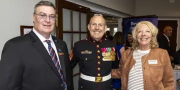 Three smiling adults at a formal event: a Marine in dress uniform with medals flanked by a man in a suit and a woman in a tan jacket nearby.