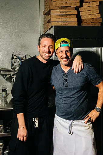 Two men in a kitchen pose for a photo; one wears a black hoodie, the other a gray shirt with a yellow cap and apron, smiling with arms around each other.