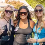 MDWineFest_featuredimage - Carroll News | Carroll Publications | Carroll County, MD Three women wearing sunglasses pose together at an outdoor festival, each holding a wine glass.