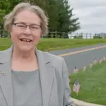 SallieTaylor_ScreenGrab - Carroll News | Carroll Publications | Carroll County, MD Smiling older woman in a gray blazer stands beside a road with a row of small American flags on the grass behind her.