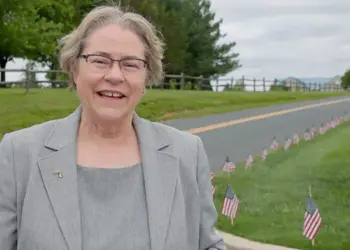 Smiling older woman in a gray blazer stands beside a road with a row of small American flags on the grass behind her.