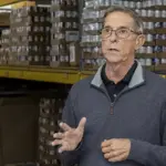Steve Lambertson Talking - Carroll News | Carroll Publications | Carroll County, MD Older man wearing glasses speaks and gesticulates in a warehouse aisle with shelves of boxed goods behind him, suggesting a briefing or tour.