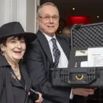 Yvonne-JP-Zeminski Winner - Carroll News | Carroll Publications | Carroll County, MD Two smiling adults pose at an event, with a man holding an open hard-shell equipment case containing papers and items for auction or raffle in a banquet setting.