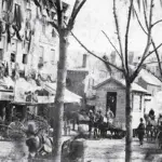 hsccmd-semicentennial-parade-Sept2024 - Carroll News | Carroll Publications | Carroll County, MD Historic city street with horse-drawn carriages, vendors, and pedestrians along a row of shops on a tree-lined avenue.