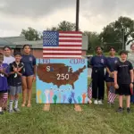 LeoPatriotism - Carroll News | Carroll Publications | Carroll County, MD Group of children and adults posing beside a handmade sign reading LEO Club USA 250, with an American flag overhead.