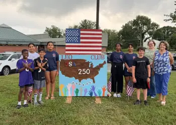 Group of children and adults posing beside a handmade sign reading LEO Club USA 250, with an American flag overhead.
