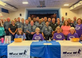 Group photo of sheriff's deputies and volunteers at a community event behind a table with The Shepherd's Staff banners in a gym setting