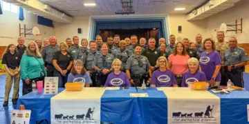 Group photo of sheriff's deputies and volunteers at a community event behind a table with The Shepherd's Staff banners in a gym setting