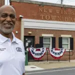 Kevin Cornick_mayor - Carroll News | Carroll Publications | Carroll County, MD Smiling man in a white polo shirt stands in front of New Windsor Town Hall with red, white, and blue bunting on the steps exterior.