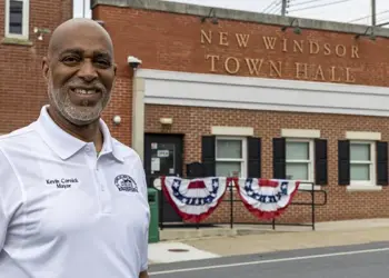Smiling man in a white polo shirt stands in front of New Windsor Town Hall with red, white, and blue bunting on the steps exterior.