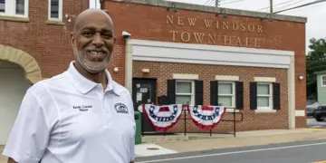 Smiling man in a white polo shirt stands in front of New Windsor Town Hall with red, white, and blue bunting on the steps exterior.
