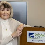 Sandy Miller - Carroll News | Carroll Publications | Carroll County, MD Smiling older woman stands at a podium with a Carroll County Workforce Development sign in a conference room.