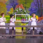 Sense-Sensibility-feature-image - Carroll News | Carroll Publications | Carroll County, MD Five women in vintage white dresses stand behind a long wooden table on a theater stage, each holding a handheld fan or tambourine, with a colorful garden backdrop and a white lattice arch centerpiece.