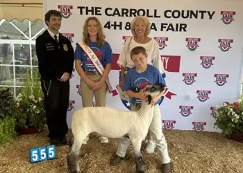 Group photo at a Carroll County 4-H Fair with a young girl wearing a sash, a boy with a sheep, a woman, and a uniformed official in front of a banner.