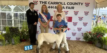 Group photo at a Carroll County 4-H Fair with a young girl wearing a sash, a boy with a sheep, a woman, and a uniformed official in front of a banner.
