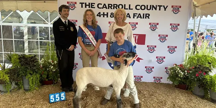 Group photo at a Carroll County 4-H Fair with a young girl wearing a sash, a boy with a sheep, a woman, and a uniformed official in front of a banner.