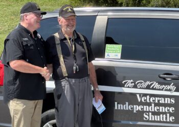 Two veterans shake hands beside a dark van with 'Veterans Independence Shuttle' lettering visible on the side.