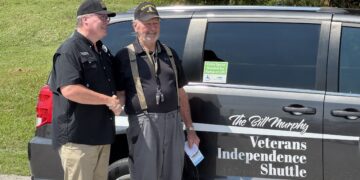 Two veterans shake hands beside a dark van with 'Veterans Independence Shuttle' lettering visible on the side.