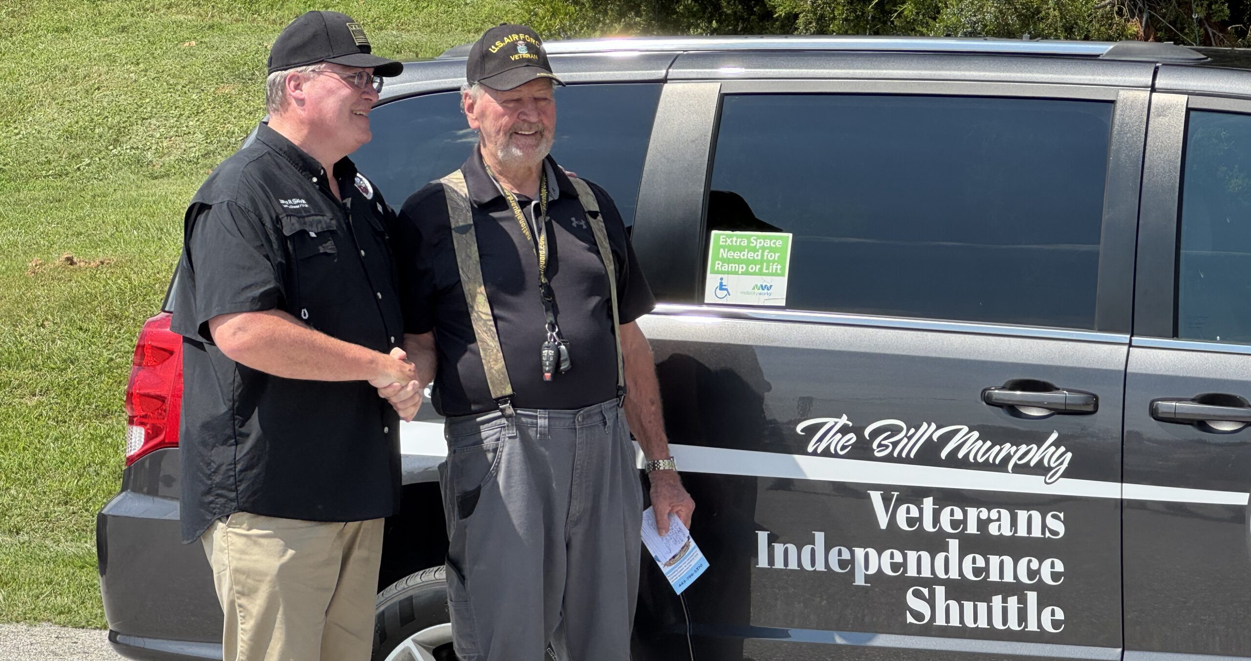 Two veterans shake hands beside a dark van with 'Veterans Independence Shuttle' lettering visible on the side.