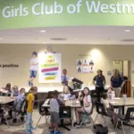 BGC_MG_9564 - Carroll News | Carroll Publications | Carroll County, MD Cafeteria scene at Boys & Girls Club of Westminster with groups of children seated at tables eating, and a few adults standing near the beige wall with posters and a large green curved sign overhead reading ’Boys & Girls Club of Westminster.’