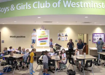 Cafeteria scene at Boys & Girls Club of Westminster with groups of children seated at tables eating, and a few adults standing near the beige wall with posters and a large green curved sign overhead reading ’Boys & Girls Club of Westminster.’