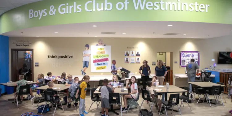 Cafeteria scene at Boys & Girls Club of Westminster with groups of children seated at tables eating, and a few adults standing near the beige wall with posters and a large green curved sign overhead reading ’Boys & Girls Club of Westminster.’