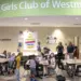 Cafeteria scene at Boys & Girls Club of Westminster with groups of children seated at tables eating, and a few adults standing near the beige wall with posters and a large green curved sign overhead reading ’Boys & Girls Club of Westminster.’